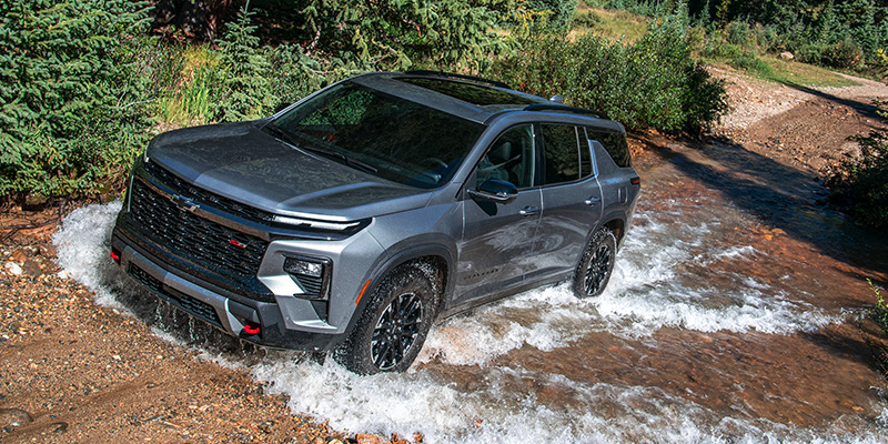 High-angle view of a gray Chevrolet Traverse splashing through a shallow, rocky stream in a wooded area.