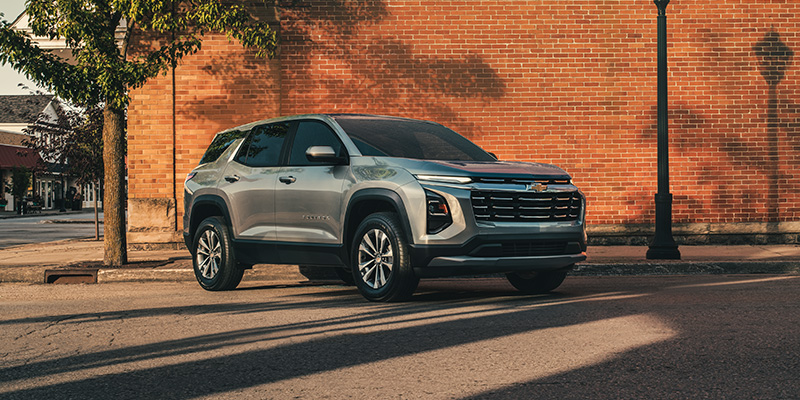 Front-three-quarter view of a silver Chevrolet Equinox parked on a town street in front of a red brick building during sunset.