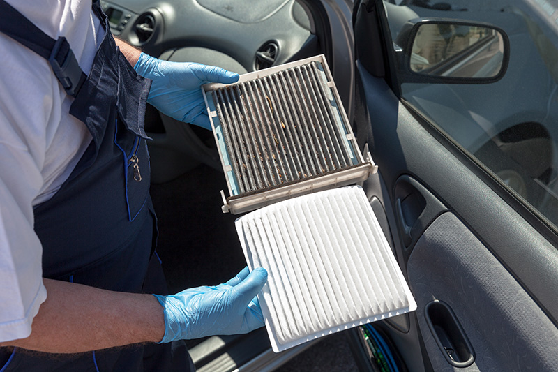 A mechanic wearing blue gloves holding a dirty, debris-filled car cabin air filter next to a clean white replacement filter outside an open car door.