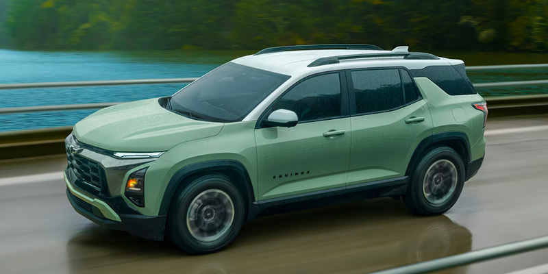 Front-three-quarter view of a sage green Chevrolet Equinox SUV with a white roof driving across a bridge on a rainy day next to a body of water.